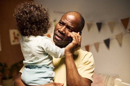 Téléchargez les photos : Sérieux père afro-américain occupé au téléphone alors qu'il était assis avec son bébé à la maison - en image libre de droit