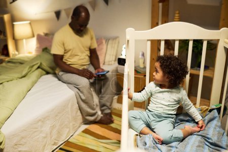Téléchargez les photos : Bébé mignon jouant dans son lit pendant que papa utilise son smartphone assis sur le lit dans la chambre - en image libre de droit