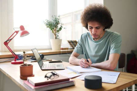 Téléchargez les photos : Jeune homme assis à table avec des gadgets et de prendre des notes dans le bloc-notes, il étudie à la maison - en image libre de droit