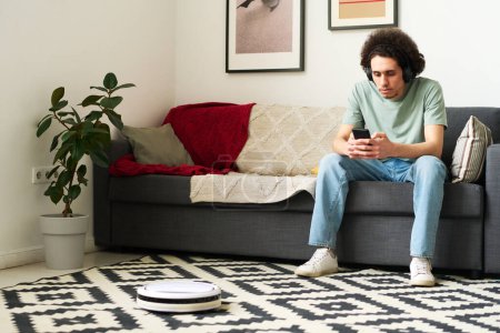 Photo for Young man in wireless headphones connecting smartphone with robot vacuum cleaner while sitting on sofa in the living room - Royalty Free Image