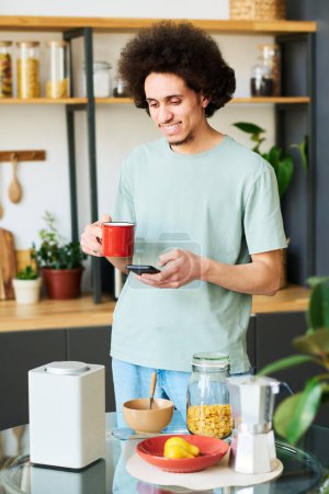 Photo for Vertical image of young man connecting smartphone with smart speaker to listen to music during his breakfast in the kitchen - Royalty Free Image