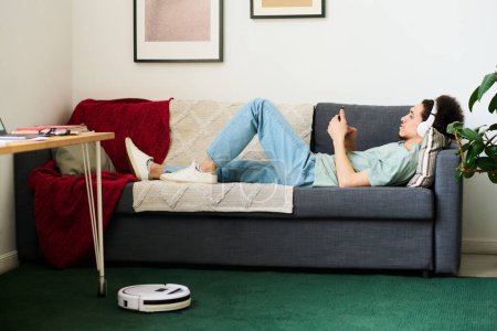 Photo for Young man in headphones lying on sofa and watching something on smartphone while robot vacuum cleaner vacuuming the carpet in the room - Royalty Free Image