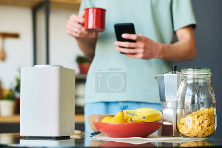 Foto de Primer plano del joven que conecta el altavoz inteligente al teléfono para escuchar música mientras está de pie en la cocina - Imagen libre de derechos