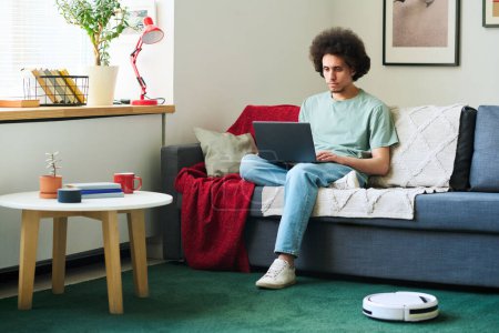 Photo for Young man sitting on sofa and using laptop while robot vacuum cleaner vacuuming the carpet in the living room - Royalty Free Image