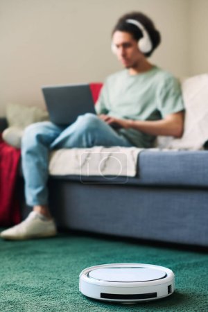 Téléchargez les photos : Image verticale du robot aspirateur nettoyant le tapis dans la pièce avec l'homme à l'aide d'un ordinateur portable assis sur le canapé en arrière-plan - en image libre de droit