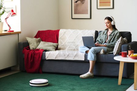 Photo for Young woman working online on laptop sitting on sofa during housework with robot vacuum cleaner - Royalty Free Image