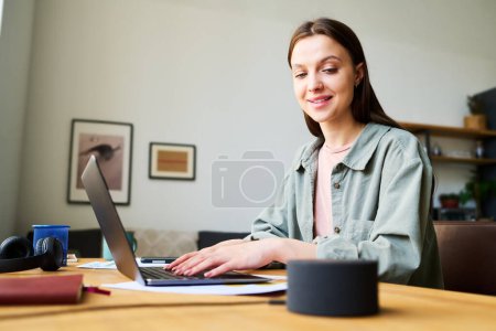 Foto de Mujer joven hablando con altavoz inteligente mientras trabaja en línea en el ordenador portátil sentado en la mesa en la habitación - Imagen libre de derechos