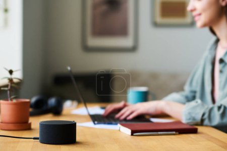 Téléchargez les photos : Gros plan du haut-parleur intelligent debout sur la table avec une femme travaillant sur un ordinateur portable en arrière-plan - en image libre de droit