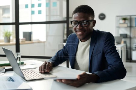 Junger, gut gelaunter Analyst oder Makler mit Brille und Formalbekleidung, der auf dem Tablet-Bildschirm online Finanzdaten durchblättert und dabei Laptop benutzt