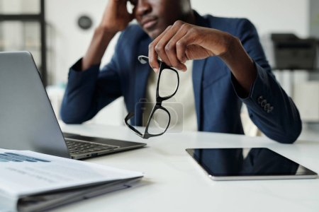 Téléchargez les photos : Focus sur la main de jeune homme d'affaires afro-américain coûteux tenant des lunettes tout en étant assis à côté du bureau avec ordinateur portable et tablette au bureau - en image libre de droit