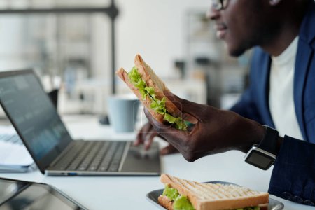 Téléchargez les photos : Focus sur sandwich végétarien appétissant en main du jeune manager afro-américain affamé en tenue de cérémonie à l'aide d'un ordinateur portable lors de la pause déjeuner - en image libre de droit