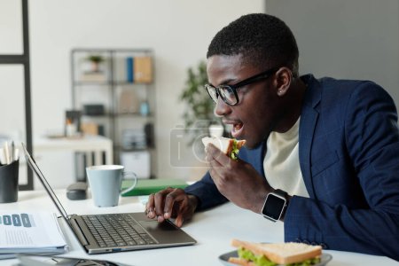 Photo for Side view of young hungry businessman eating sandwich in front of laptop screen by workplace while watching online video or organizing work - Royalty Free Image