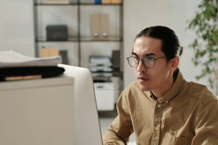 Téléchargez les photos : Jeune analyste sérieux en lunettes et chemise beige assis par le lieu de travail devant l'ordinateur et regardant à travers les données en ligne à l'écran - en image libre de droit