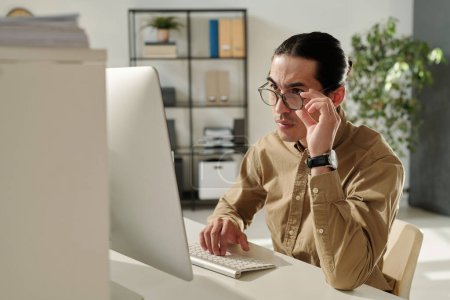 Téléchargez les photos : Jeune chef de la direction ou directeur financier confiant regardant l'écran d'ordinateur à travers des lunettes tout en organisant le travail - en image libre de droit