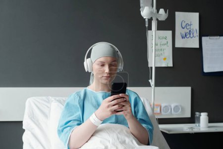 Mujer joven seria con auriculares viendo videos en línea o mensajes de texto en un teléfono inteligente mientras está sentada en la cama en la sala del hospital después del tratamiento médico