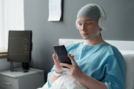 Foto de Paciente joven del departamento oncológico de clínicas que usa smartphone mientras descansa en la cama después del tratamiento médico - Imagen libre de derechos
