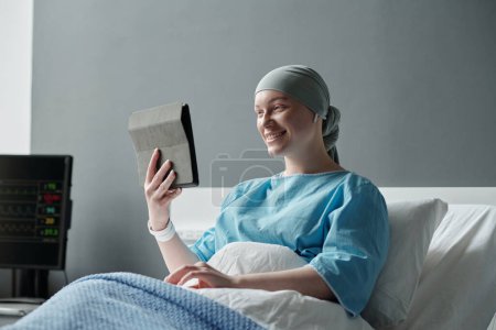 Photo for Young smiling woman with oncological illness looking at tablet screen while communicating with her family in video chat in hospital ward - Royalty Free Image