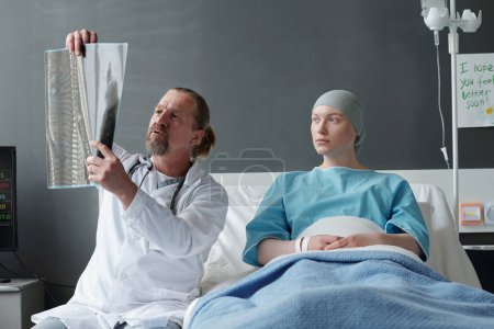 Foto de Paciente joven con pañuelo en la cabeza escuchando a su médico comentando la imagen de rayos X del cerebro mientras está sentada en la cama junto a ella en la sala del hospital - Imagen libre de derechos