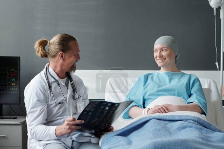Foto de Mujer joven y feliz con pañuelo en la cabeza sentada en la cama y hablando con su médico con resultados de resonancia magnética cerebral durante el curso de tratamiento médico - Imagen libre de derechos