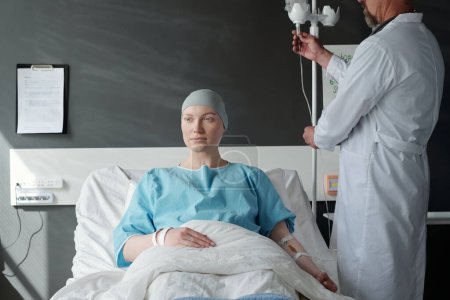 Foto de Mujer joven enferma con cáncer sentada en la cama durante el tratamiento de quimioterapia mientras es oncóloga masculina con bata de laboratorio parada junto a ella - Imagen libre de derechos