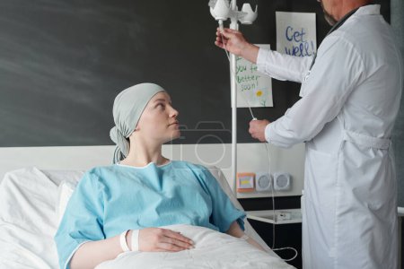 Paciente joven con cáncer sentada en la cama y mirando al oncólogo preparando gotero para quimioterapia mientras está de pie junto a la mujer