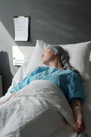 Foto de Mujer joven con pañuelo en la cabeza y camisa azul acostada en la cama en clínicas y durmiendo durante el procedimiento médico mientras recibe tratamiento de quimioterapia - Imagen libre de derechos