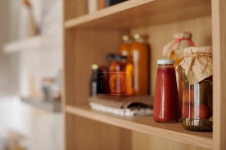 Photo for Focus on group of jars with homemade ketchup, pickled tomatoes and other food products standing on wooden shelf in the kitchen - Royalty Free Image