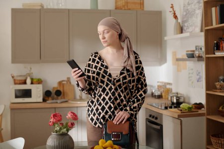Foto de Mujer joven con pañuelo en la cabeza desplazándose en el teléfono móvil mientras está de pie junto a la mesa redonda de vidrio en la cocina después de volver a casa desde el hospital - Imagen libre de derechos