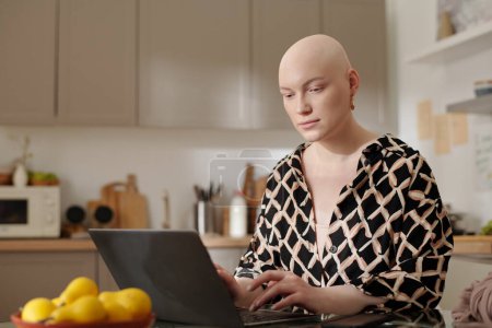 Foto de Mujer joven con la cabeza calva después de la quimioterapia escribiendo en el teclado del ordenador portátil mientras está sentado junto a la mesa delante de la cámara en la cocina - Imagen libre de derechos