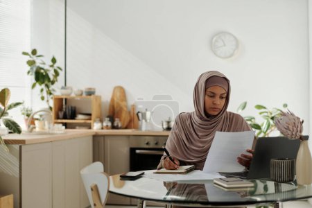 Photo for Young female employee reading financial document or contract and making notes while working over business project in home office - Royalty Free Image