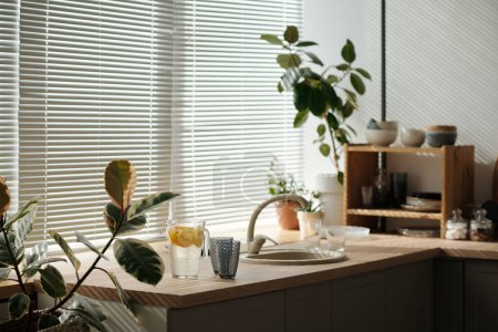 Photo for Part of kitchen counter with sink, homemade lemonade, two glasses and green domestic plants against large window with venetian blinds - Royalty Free Image
