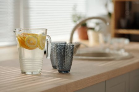 Téléchargez les photos : Pot transparent avec limonade fraîche maison rafraîchissante et deux verres debout sur le comptoir contre l'évier et une partie de la fenêtre dans la cuisine - en image libre de droit