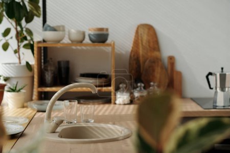 Foto de Parte del mostrador de cocina con fregadero beige y grifo para lavar platos, productos y manos contra estantes y utensilios de cocina de pared blanca - Imagen libre de derechos