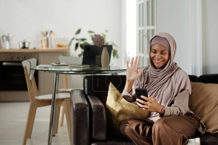 Foto de Feliz joven musulmana saludando de la mano a su amigo o marido en la pantalla del teléfono inteligente mientras se comunica en el chat de vídeo en casa - Imagen libre de derechos