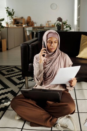 Foto de Joven mujer afroamericana en hijab hablando con el cliente en el teléfono móvil mientras está sentada en el suelo y leyendo los términos y condiciones del contrato - Imagen libre de derechos