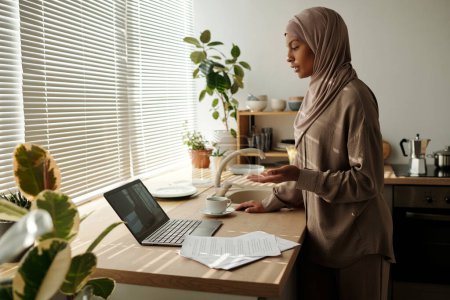 Photo for Young confident businesswoman in hijab looking at laptop screen during talk with colleague at digital meeting while explaining something - Royalty Free Image