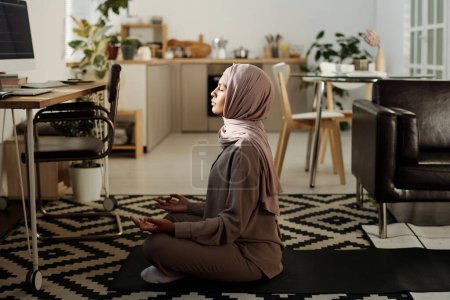 Photo for Young serene woman in hijab and jumpsuit sitting in pose of lotus on mat in front of workplace with computer monitor and meditating - Royalty Free Image