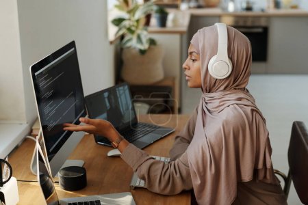 Junge muslimische Programmiererin zeigt auf Daten auf dem Computerbildschirm, während sie am Arbeitsplatz sitzt und im Videochat mit einem Kollegen kommuniziert