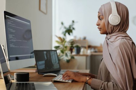 Foto de Vista lateral de la joven programadora en auriculares y hijab decodificación de datos en línea mientras se comunica en chat de vídeo con su colega - Imagen libre de derechos