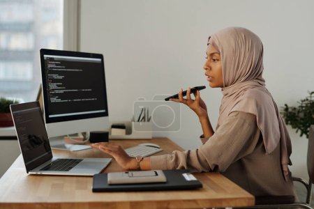 Foto de Vista lateral de la confiada programadora musulmana hablando por altavoz durante el trabajo mientras sostiene el teléfono móvil por la boca - Imagen libre de derechos