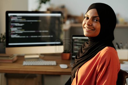 Téléchargez les photos : Jeune femme musulmane joyeuse employée en hijab et chemise rouge vous regardant tout en se tenant devant la caméra dans le bureau à la maison contre ordinateur - en image libre de droit