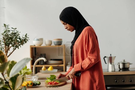Téléchargez les photos : Jeune femme en foulard noir et chemise rouge coupant des légumes frais sur planche à découper pendant la cuisson de la salade pour le petit déjeuner par comptoir de cuisine - en image libre de droit