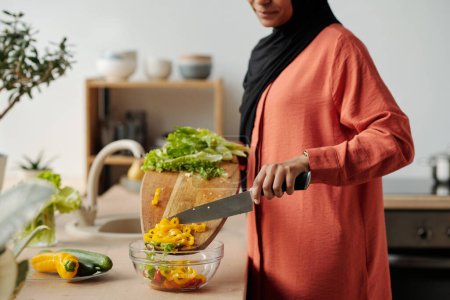 Foto de Foto recortada de ama de casa joven de pie junto al mostrador de la cocina y poner pimienta picada en un tazón mientras se prepara la ensalada de verduras para el desayuno - Imagen libre de derechos