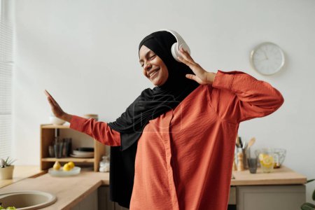 Foto de Joven mujer musulmana alegre con auriculares, hijab y camisa escuchando su música favorita, bailando y divirtiéndose delante de la cámara - Imagen libre de derechos