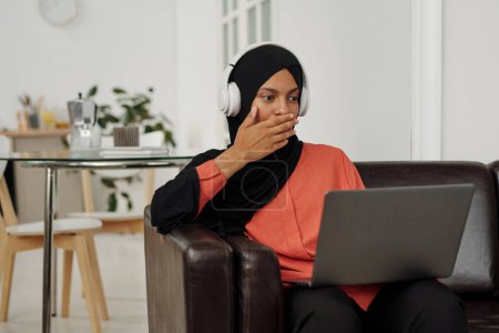 Téléchargez les photos : Jeune femme musulmane choquée dans le hijab couvrant sa bouche à la main tout en regardant une vidéo ou un film en ligne sur l'écran de l'ordinateur portable à loisir - en image libre de droit