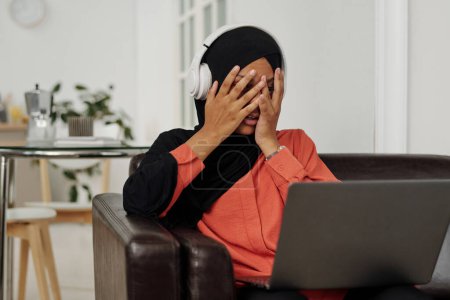 Téléchargez les photos : Jeune femme musulmane effrayée avec un ordinateur portable sur les genoux couvrant son visage avec les mains alors qu'elle était assise sur le canapé et regardant un film d'horreur ou un thriller - en image libre de droit