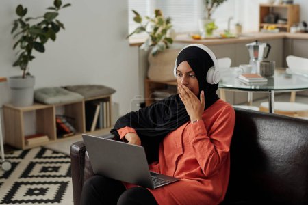 Téléchargez les photos : Jeune femme terrifiée ou surprise, musulmane couvrant la bouche avec la main tout en regardant l'écran de l'ordinateur portable et regarder thriller ou vidéo terrifiante - en image libre de droit