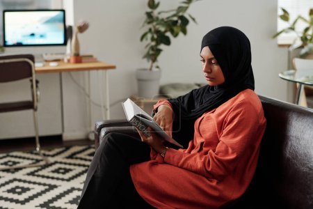 Foto de Joven estudiante musulmana en hijab y ropa casual leyendo enciclopedia psicología mientras revisa asignación a domicilio o se prepara para el examen - Imagen libre de derechos