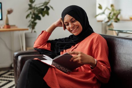 Foto de Feliz joven musulmana en hiyab leyendo artículo psicológico en enciclopedia mientras se sienta en el sofá en casa y se prepara para el examen - Imagen libre de derechos