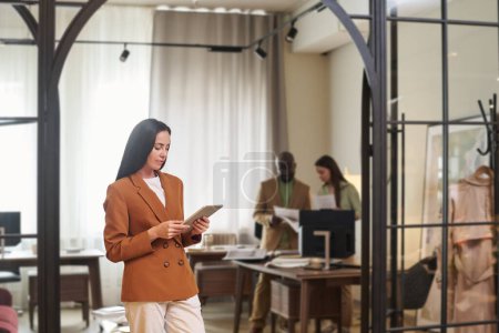 Foto de Joven diseñadora femenina seria mirando la pantalla de la tableta mientras está de pie frente a la cámara en el área de coworking y desplazándose a través de datos en línea - Imagen libre de derechos